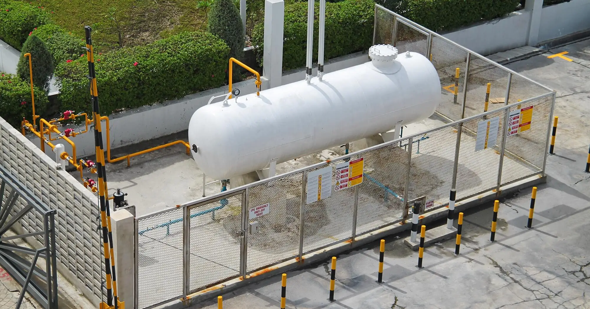 Outdoor LPG storage tank enclosed by a safety fence with warning signs and protective barriers.
